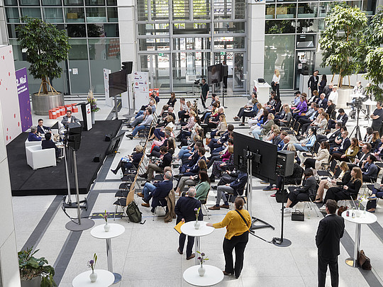 Blick von oben in einen hellen Raum mit Bühne und Publikum, was davor sitzt. Auf der Bühne sind Sessel und ein Rednerpult zu sehen. 