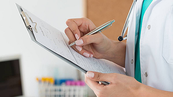 Ein medizinischer Mitarbeiter im weißen Kittel hält ein Klemmbrett und einen Stift in der Hand und schreibt auf ein Formular. Um seinen Hals hängt ein Stethoskop. Im Hintergrund sind verschwommene medizinische Hilfsmittel und ein Computer zu sehen.