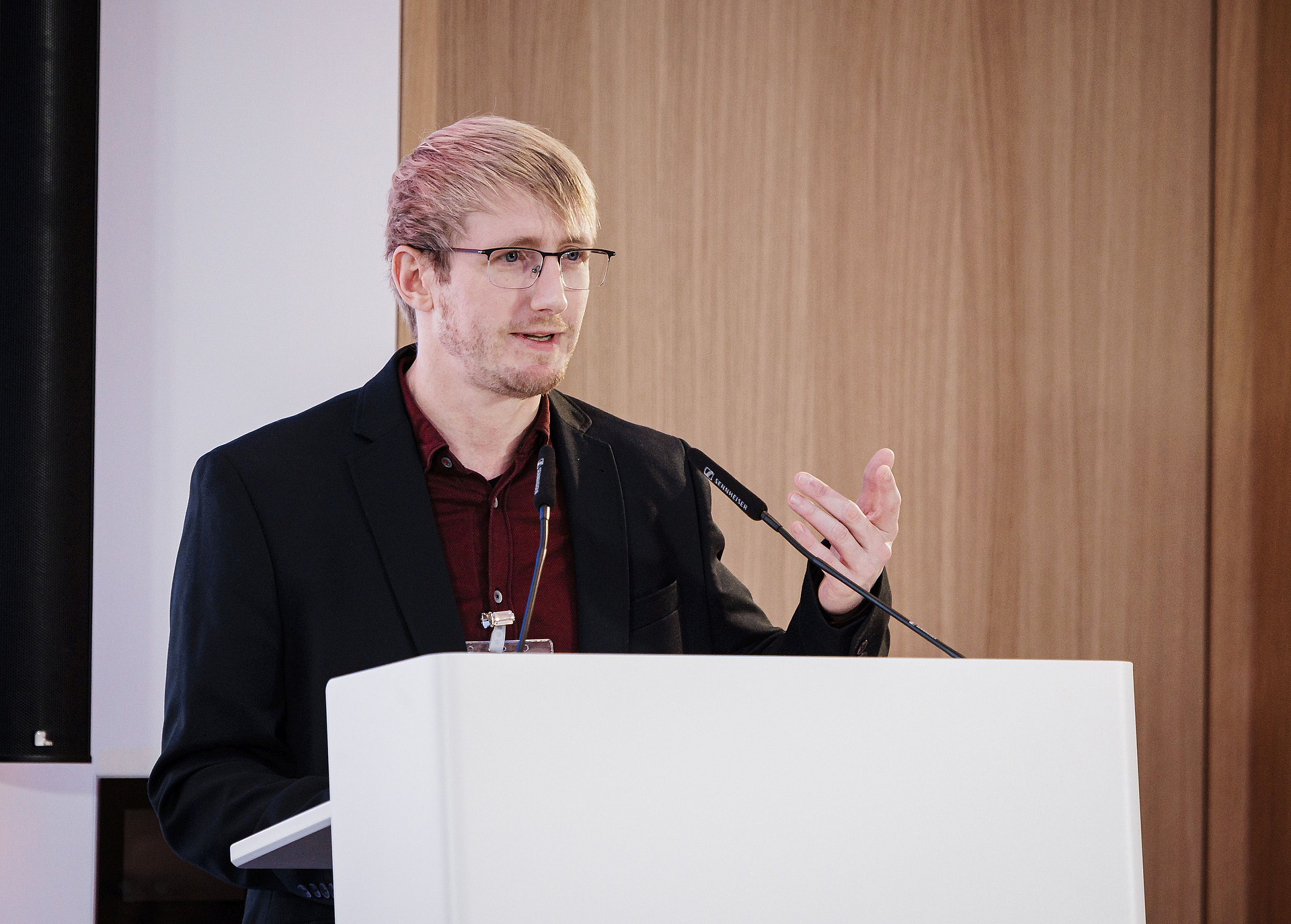 Eine Person mit hellem Haar, Brille und rotem Hemd spricht an einem Podium. Sie gestikuliert mit einer Hand, vor ihr steht ein Mikrofon. Im Hintergrund ist eine Holzpaneelwand.