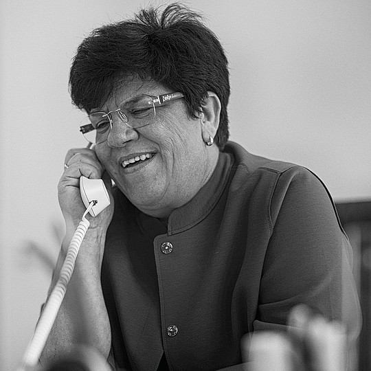 Schwarz-Weiß-Foto: Claudia Moll sitzt lächelnd an einem Schreibtisch und hält sich einen Telefonhörer ans Ohr.