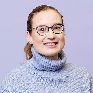 Eine Frau mit Brille und zurückgebundenem hellbraunem Haar lächelt in die Kamera. Sie trägt einen hellblauen, gerippten Rollkragenpullover vor einem schlichten lavendelfarbenen Hintergrund.