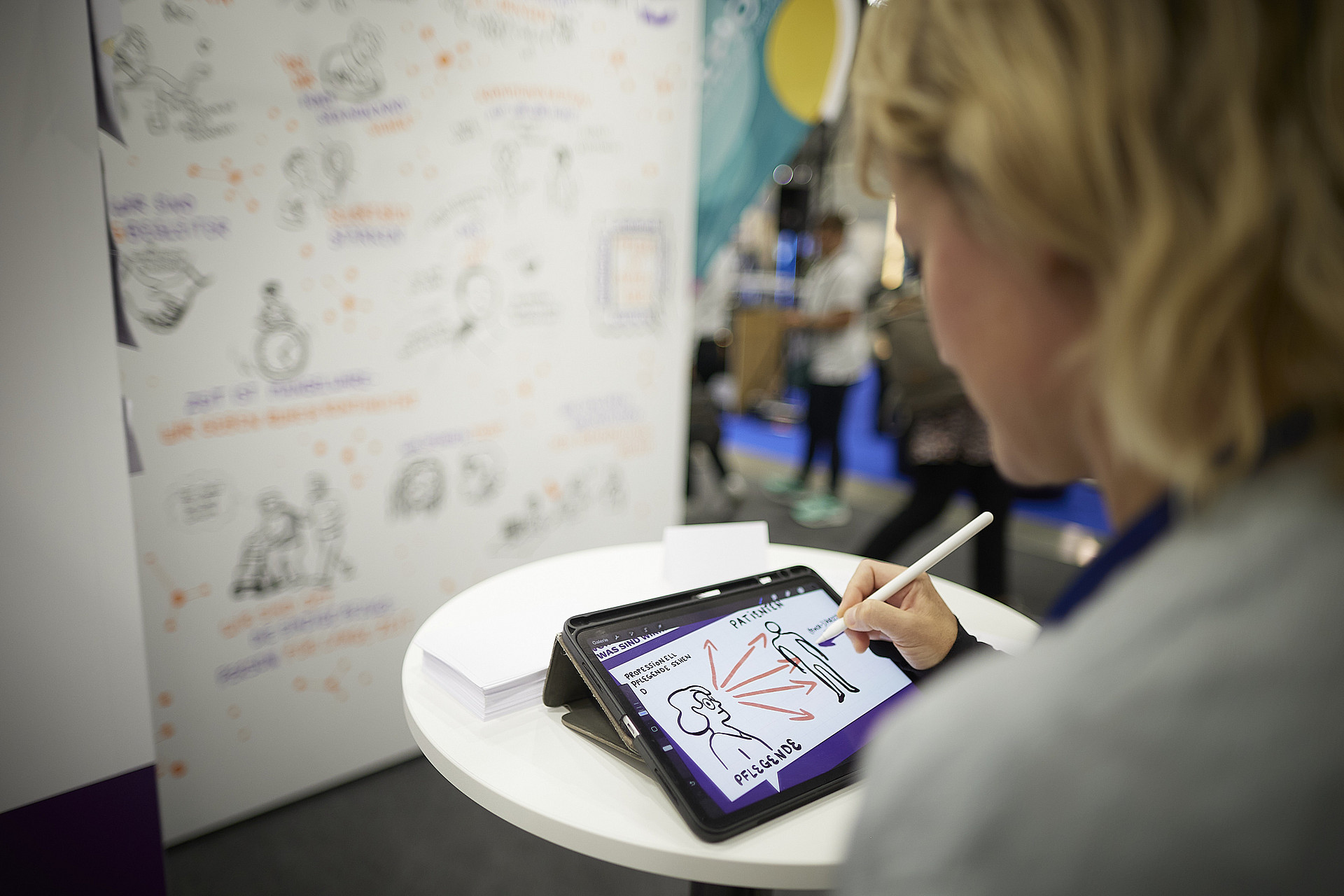 Eine Person verwendet ein Tablet mit einem Stift, um Figuren zu zeichnen. Auf dem Bildschirm sind zwei umrissene Figuren mit Text zu sehen. Im Hintergrund sind auf einem Whiteboard verschiedene Abbildungen und Notizen zu sehen. Das Setting scheint eine Veranstaltung oder ein Workshop zu sein.