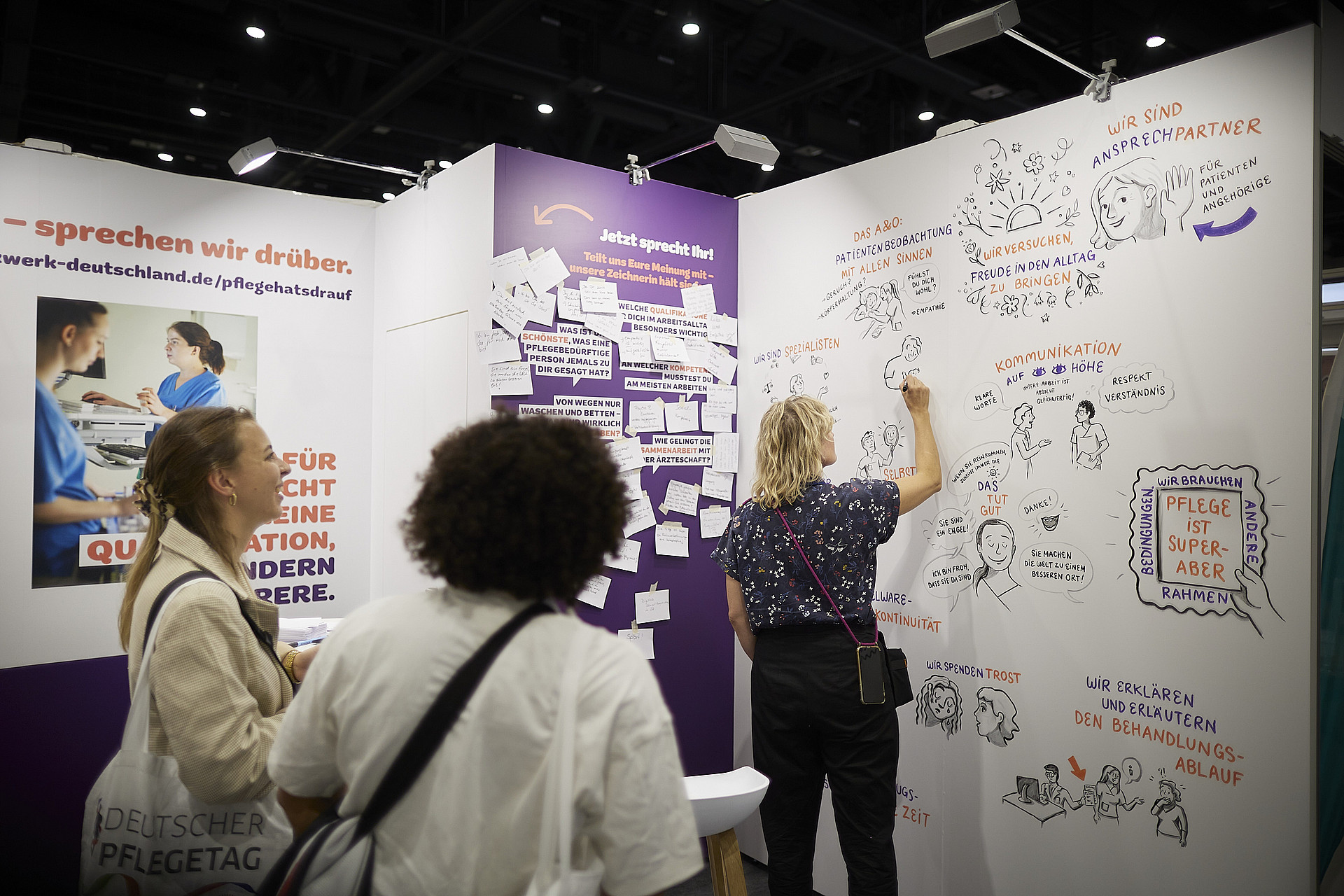 Menschen an einem Messestand interagieren mit einer Wand, auf der illustrierte Notizen zu sehen sind. Eine Person schreibt an die Wand. Die Notizen und Illustrationen konzentrieren sich auf Kommunikation und Partnerschaften. Im Hintergrund sind Banner in deutscher Sprache zu sehen.