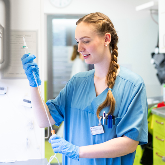 Eine Mitarbeiterin des Gesundheitswesens in blauer Uniform und Handschuhen justiert medizinische Geräte in einer hell erleuchteten Klinikumgebung. Sie trägt einen Zopf und einen Ausweis. Im Hintergrund sind verschiedene medizinische Hilfsmittel zu sehen.
