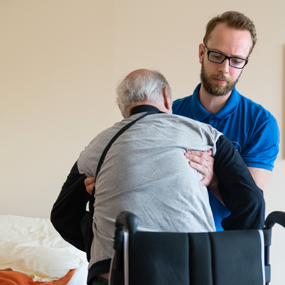 Eine Pflegekraft mit Brille und blauem Hemd hilft einem älteren Mann mit grauem Haar und grauem Hemd, aus dem Rollstuhl zu steigen. Sie sind im Haus, im Hintergrund ist ein Bett zu sehen.