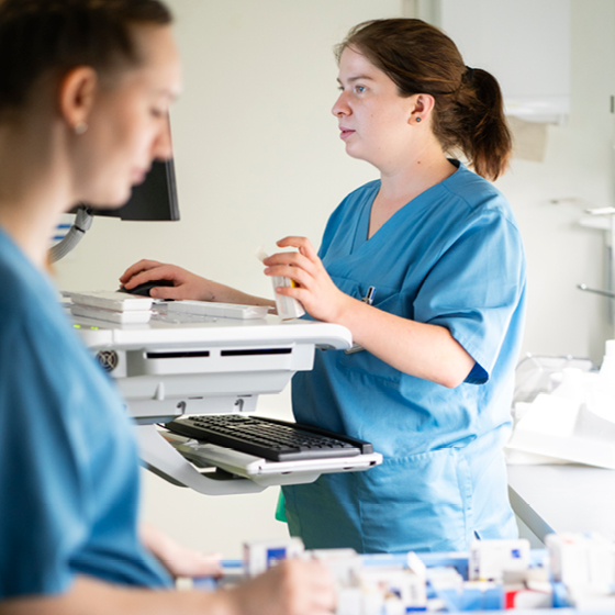 Zwei medizinische Fachkräfte in blauen OP-Kitteln arbeiten in einem Krankenhaus. Einer konzentriert sich auf einen Computer, während der andere im Vordergrund medizinisches Material handhabt. Der Raum ist hell und mit medizinischen Geräten ausgestattet.