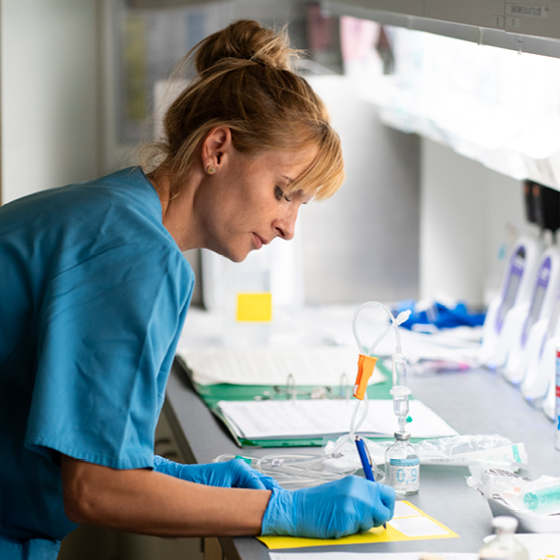 Eine Person in einem blauen OP-Kittel und Handschuhen arbeitet in einem Labor. Sie schreibt auf einem Blatt auf einer Theke, die mit Laborgeräten, darunter einer Pipette und Behältern, gefüllt ist. Im Hintergrund sind Regale mit Gegenständen zu sehen.