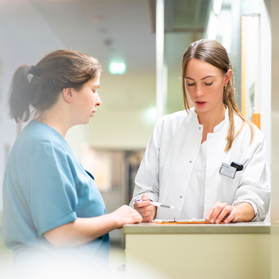 Zwei medizinische Fachkräfte besprechen in einem Krankenhausflur Notizen auf einem Klemmbrett. Einer trägt eine blaue Uniform, der andere einen weißen Kittel und hält einen Stift in der Hand. Die Szene ist hell beleuchtet und der Hintergrund ist unscharf.