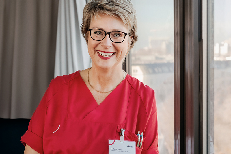 Eine lächelnde Frau mit kurzen blonden Haaren und Brille, die einen roten Kittel und ein Namensschild trägt, steht in einem Gebäude neben einem Fenster. Im Hintergrund sind Stadtgebäude zu sehen.