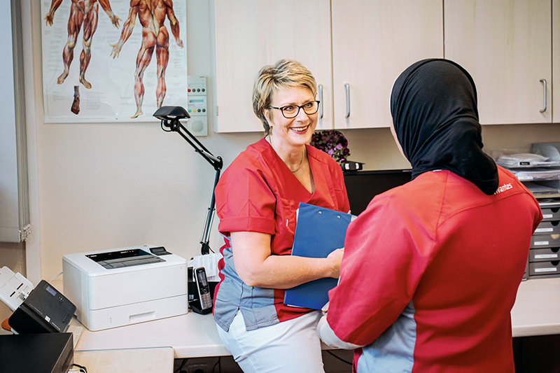 Zwei medizinische Fachkräfte in roten Uniformen unterhalten sich in einer Arztpraxis. Die eine sitzt lächelnd und hält ein Klemmbrett in der Hand; die andere steht mit dem Rücken zur Kamera und trägt ein schwarzes Kopftuch. An der Wand hängt eine Muskelanatomietafel.