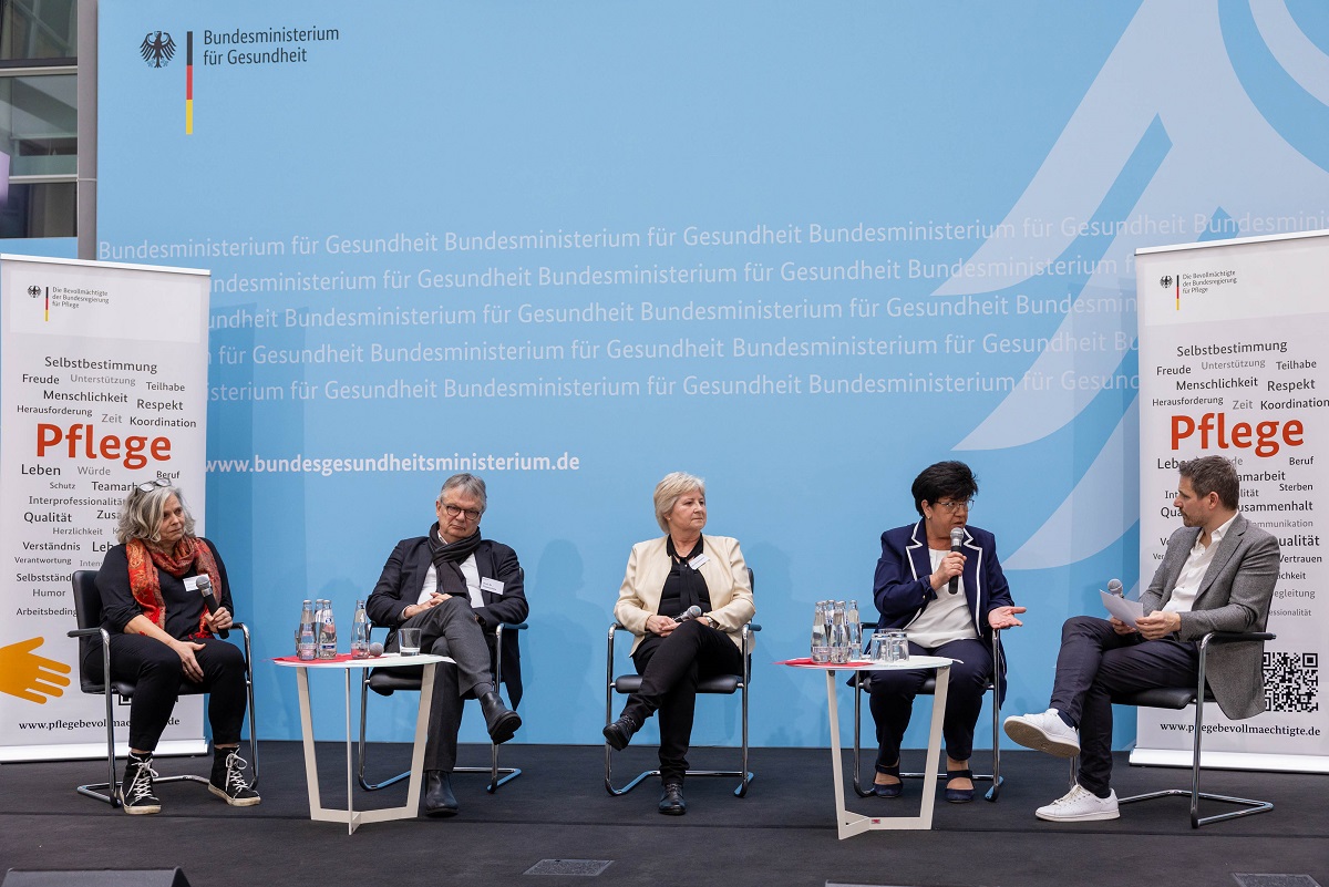 Fünf Personen sitzen mit Mikrofonen auf einer Bühne und nehmen an einer Podiumsdiskussion teil. Im Hintergrund ist das Logo des Bundesministeriums für Gesundheit zu sehen. Auf der Bühne sind Banner mit Hinweisen auf das Gesundheitswesen zu sehen.