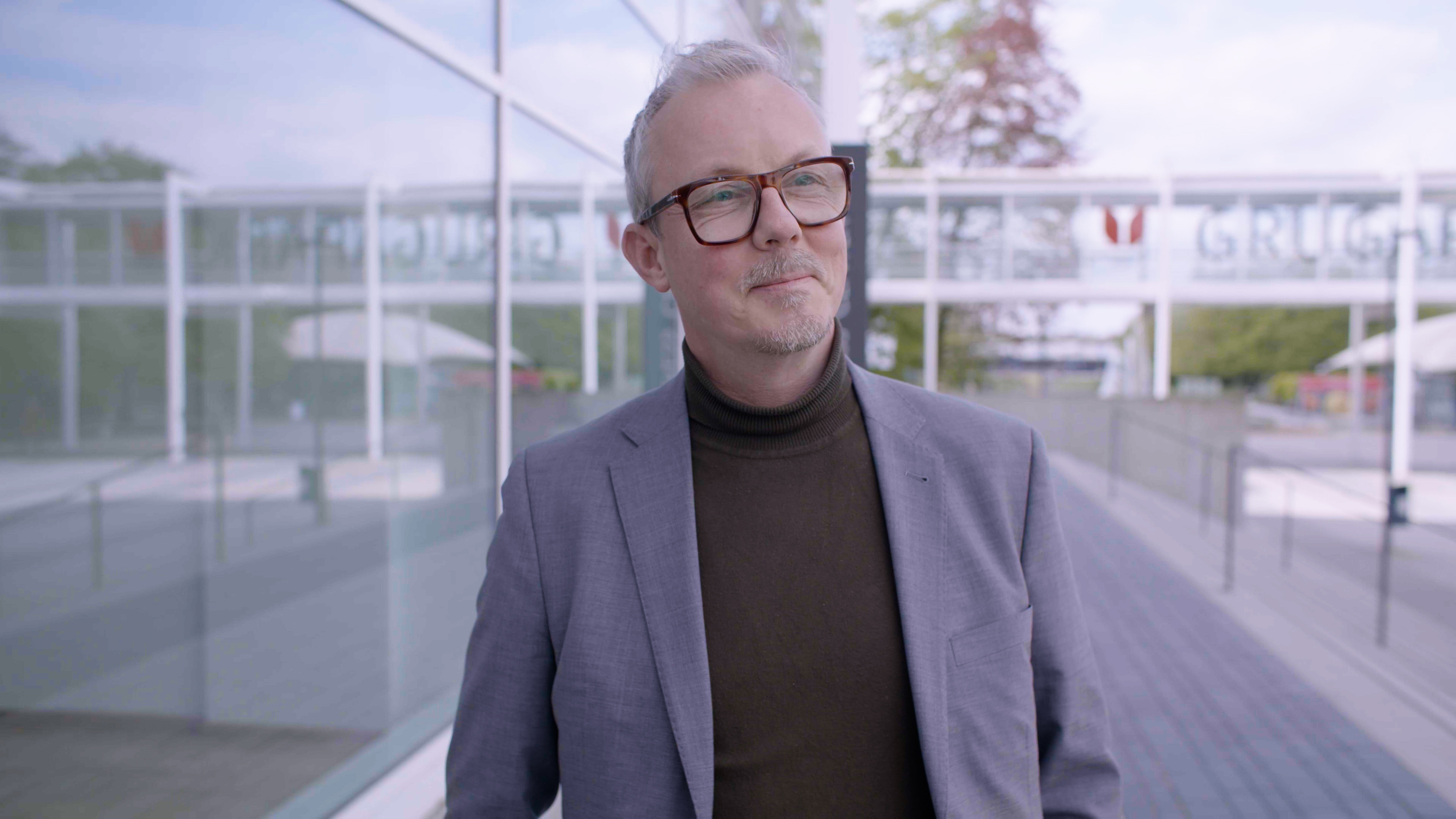 Ein Herr mit grauem kurzem Haar und Brille ist vor einen Glasgebäude zu sehen. Er schaut zur Kamera. Es ist Prof. Isfort.